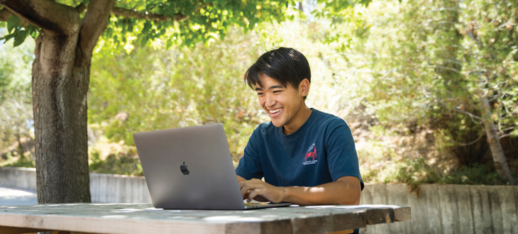 student sits at a picnic table, looking at his Macbook screen