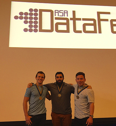 Three men with medals around their necks pose in front of a projector that reads ASA DataDest