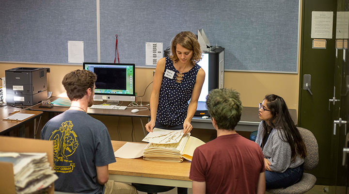 Katie Pearson picks up a piece of paper with a dried plant on it from a stack of more paper and plants. Three students watch her.