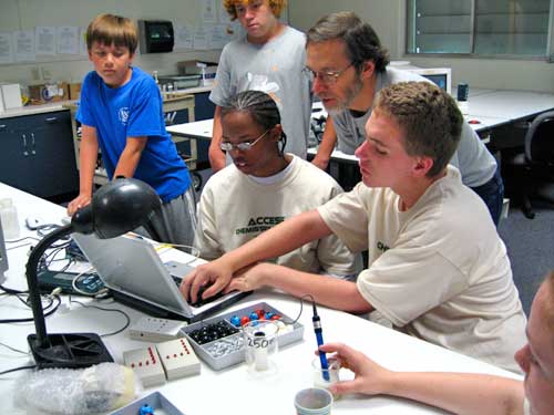 Students and instructor gathered around laptop in chemistry lab.