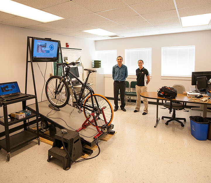 Robert Clark and Christiane O’Hara stand next to a bicycle set up to a monitor