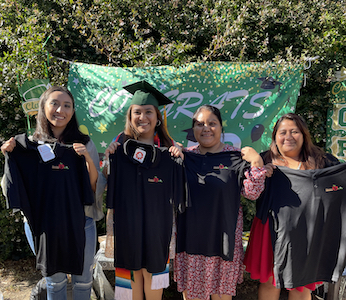 Four students, one in graduation robes, holding up black polo shirts in front of a Congrats sign