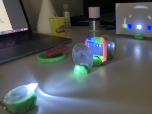 an led light shining through a lens, a gradient, another lens, and onto a piece of white paper, all set in playdough. White light, a spectrum, and shadows show on the paper.