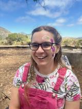 Picture of a white woman wearing glasses, pink overalls, and a floral t-shirt. Her face is painted with purple flowers. 