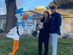 Alex Herrington, his partner, and their baby standing in front of their house by a stork cutout that says "Welcome Graham!"