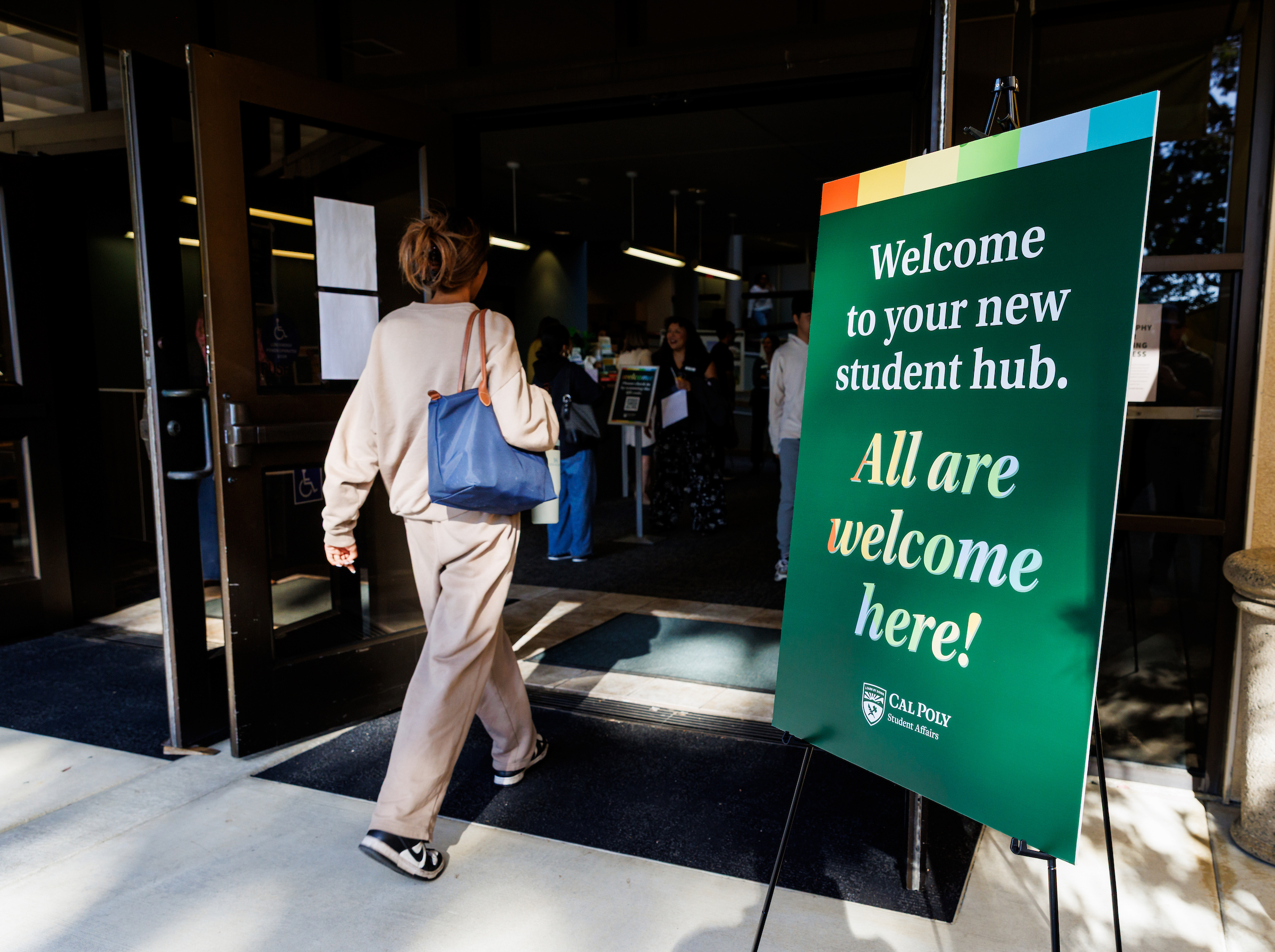 A woman walks into Building 15 during a welcome event in October 2025. A sign reads, "Welcome to your new student hub. All are welcome here!"
